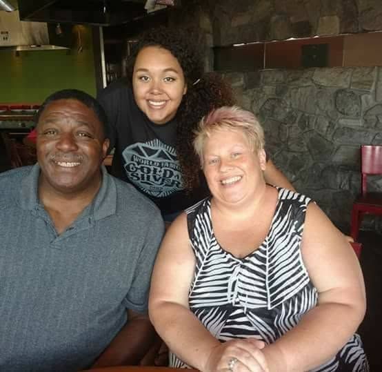 Three smiling people seated together in a dim restaurant booth, with one standing behind them.