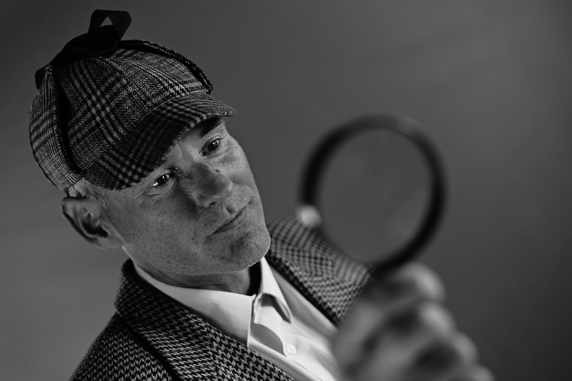 A greyscale picture of an old man with a hat, holding a magnifying glass.