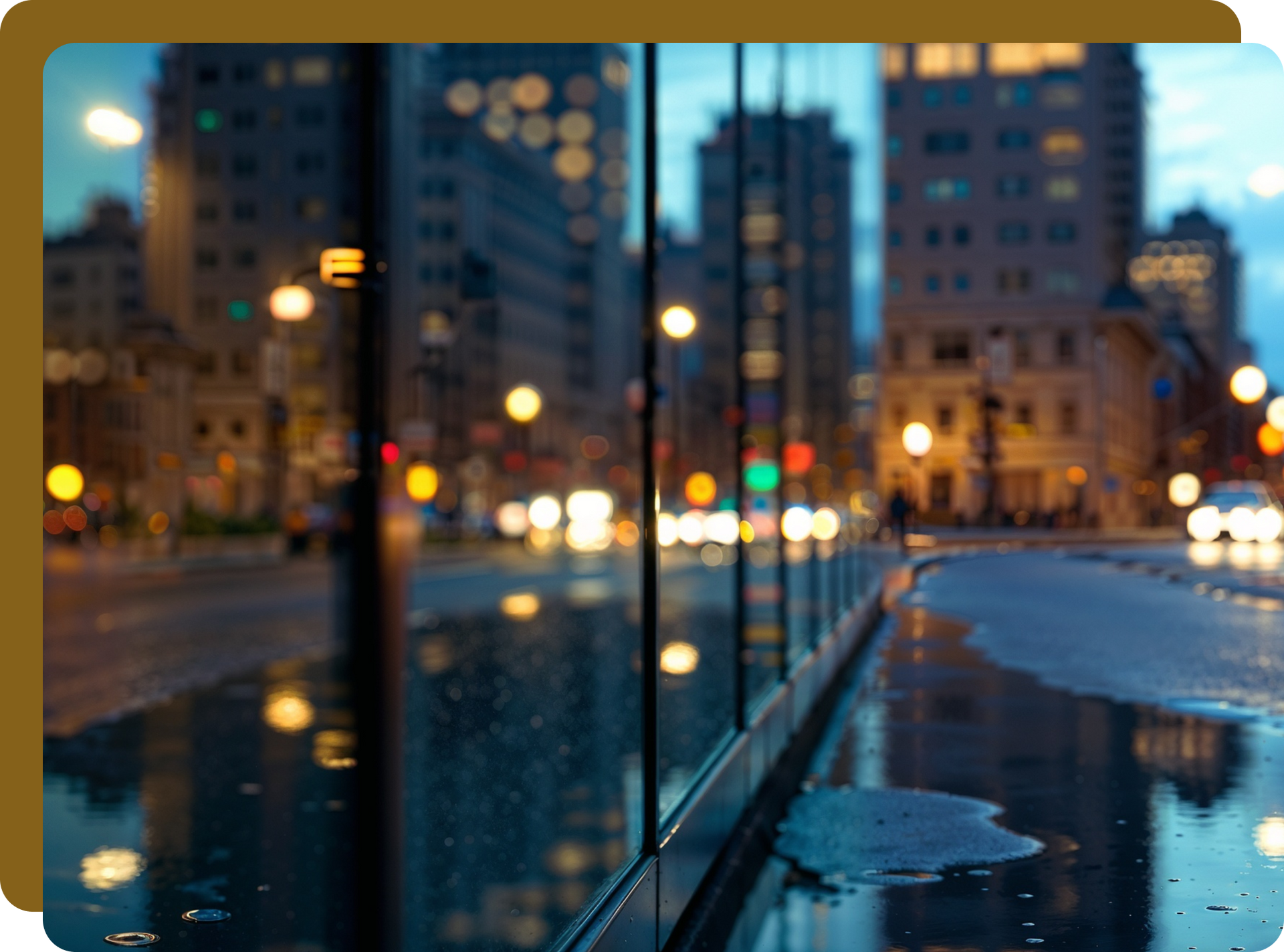 City street at dusk, lights reflected in wet sidewalk. Buildings in background, glass wall in foreground.