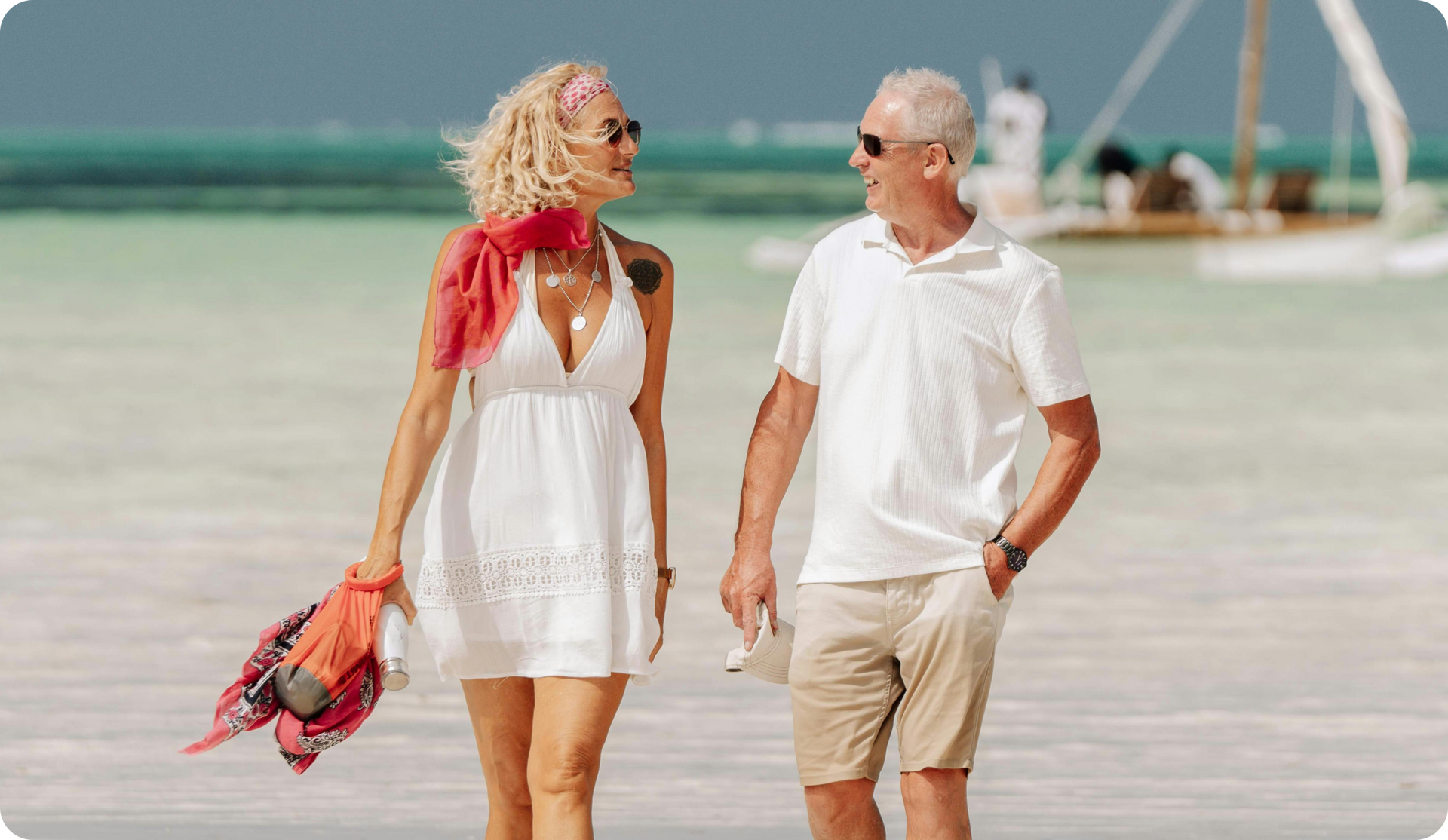 Couple strolling on a white sand beach, turquoise water, woman in white dress, man in khaki shorts, smiling, sunglasses.