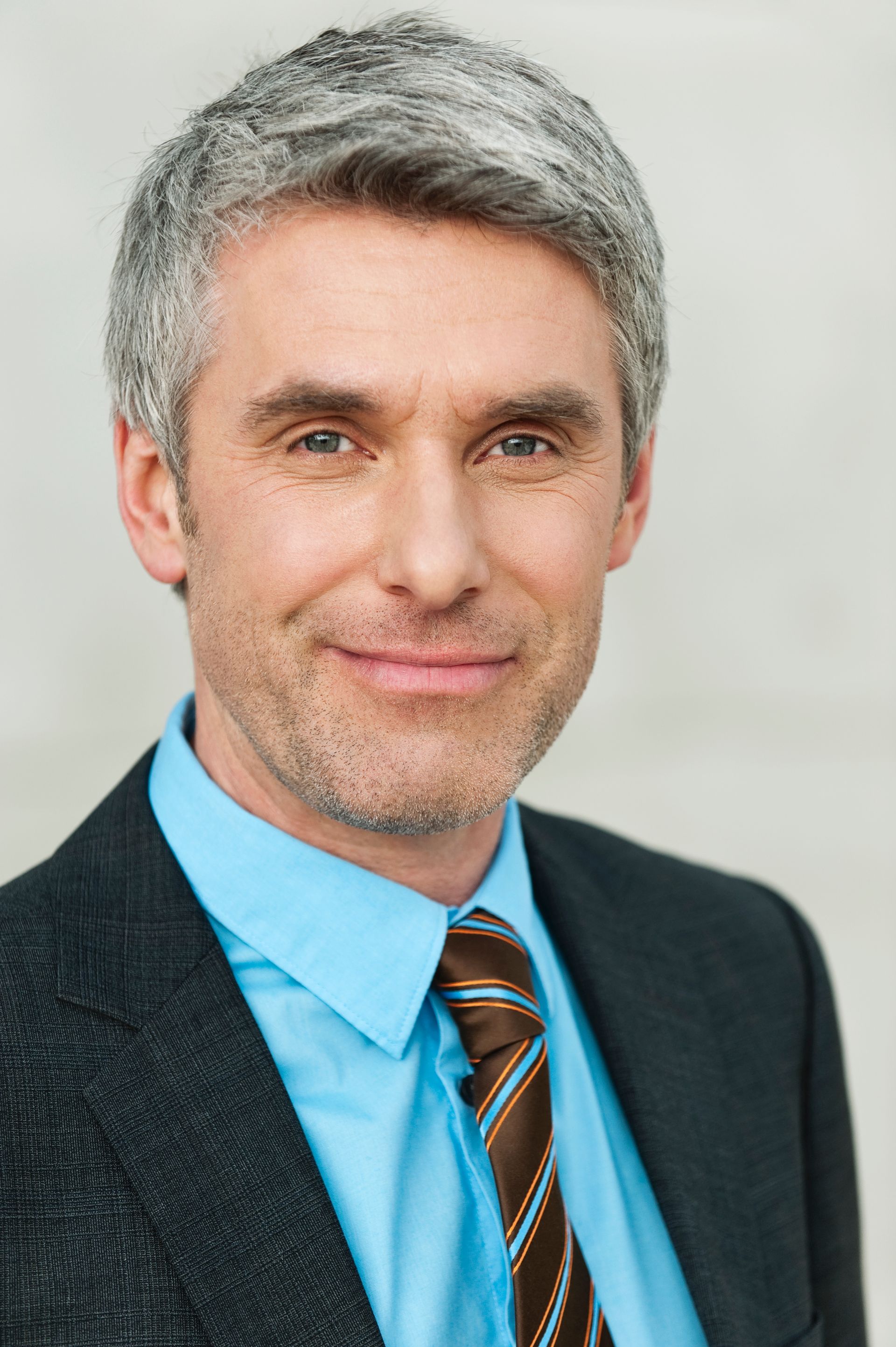 Professional headshot of a man in a blue shirt and striped tie, wearing a dark blazer, smiling at the camera