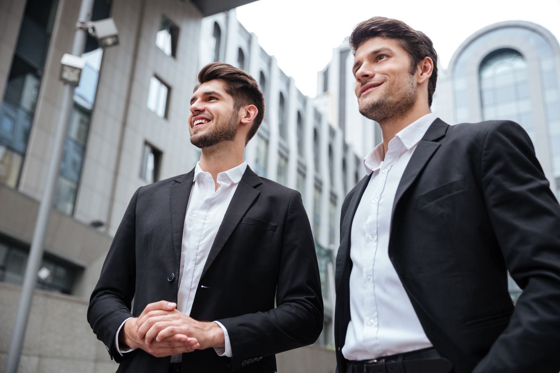 Two professionals in dark suits standing outdoors beside office buildings, smiling and looking ahead