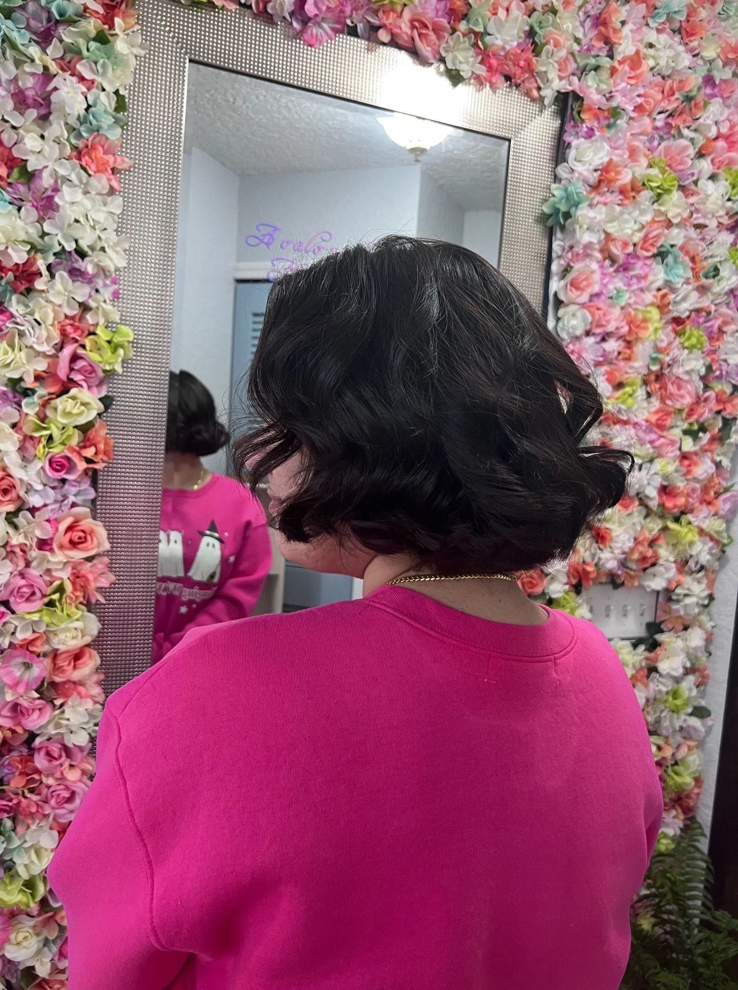 A woman in a pink sweater is standing in front of a mirror in front of a wall of flowers.