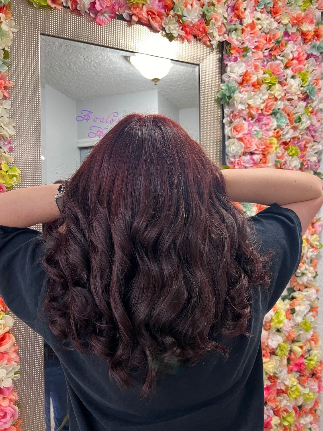 A woman is standing in front of a mirror with flowers on the wall behind her.