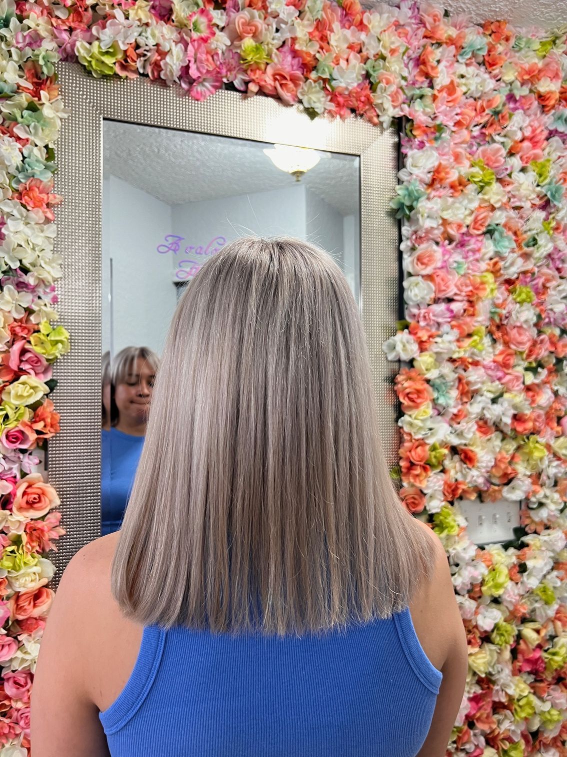 A woman is standing in front of a mirror with flowers on the wall behind her.