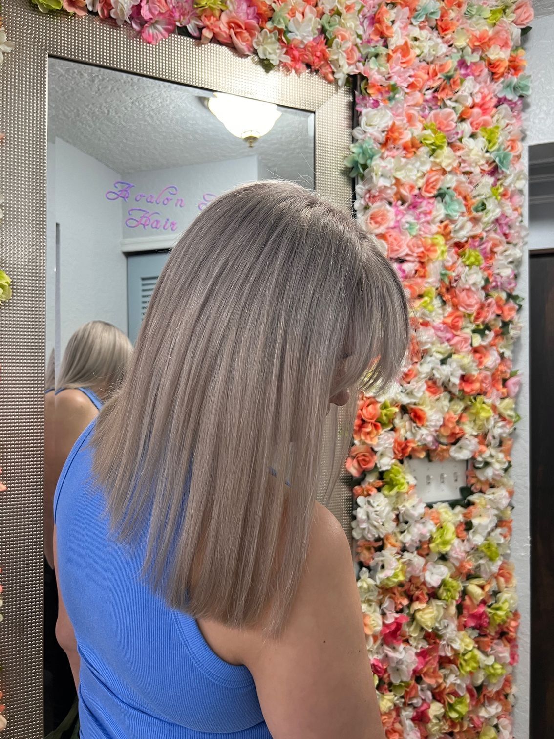 A woman is standing in front of a mirror in front of a wall of flowers.