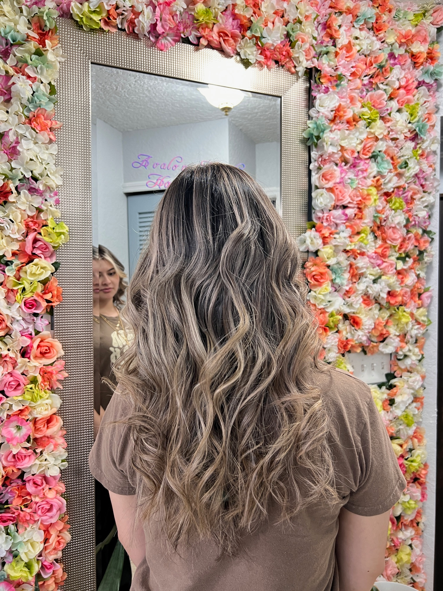 A woman is standing in front of a mirror in front of a wall of flowers.