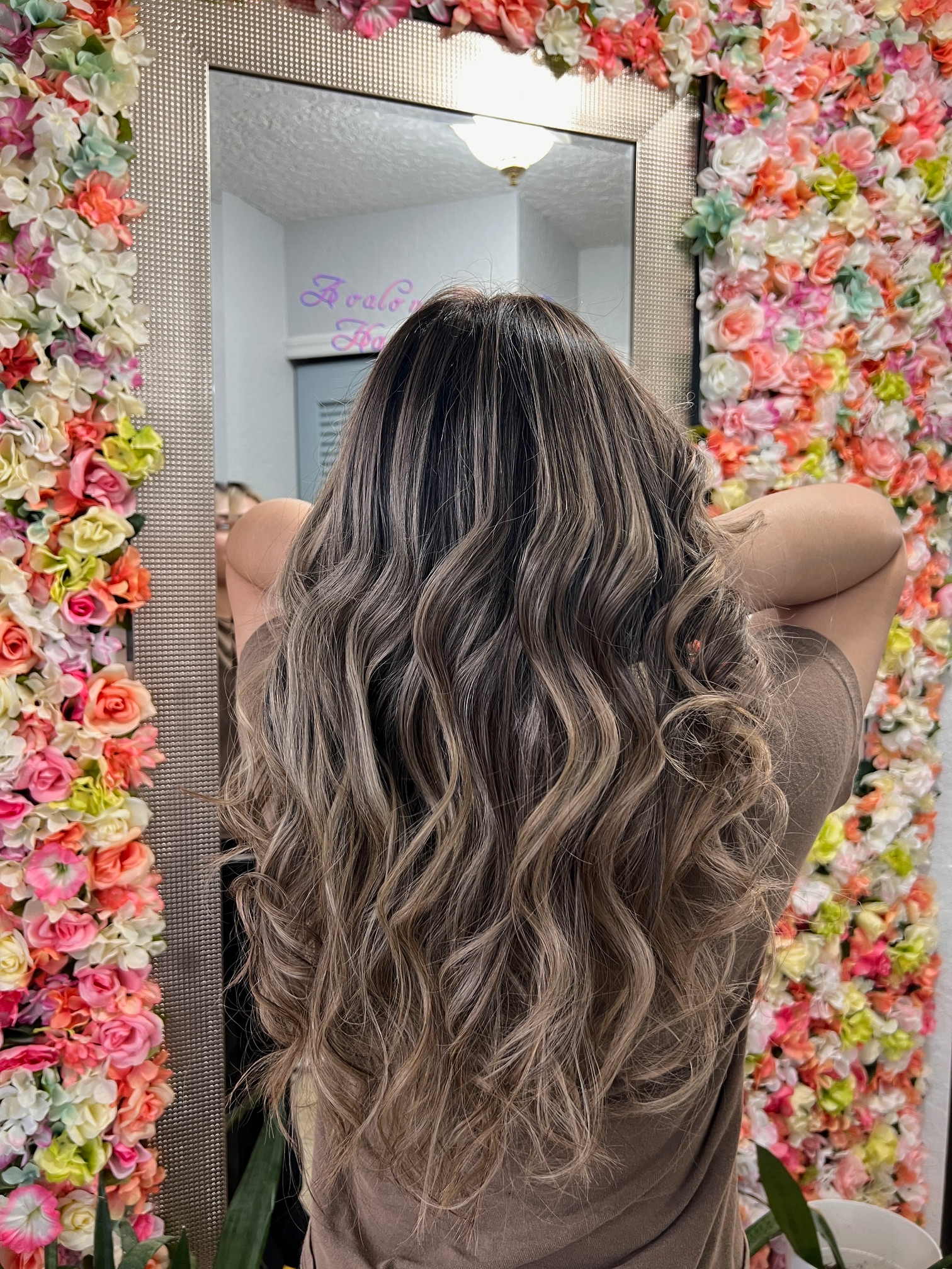 A woman is standing in front of a mirror with flowers on the wall behind her.