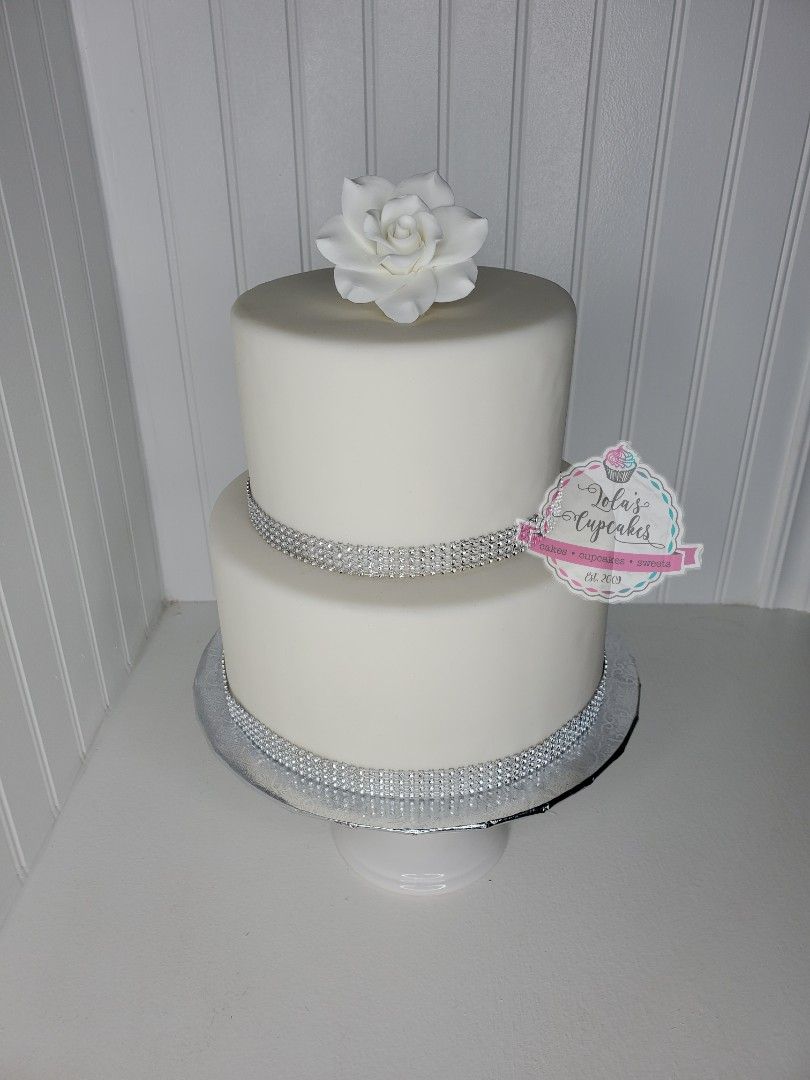 Two-tiered white cake with silver ribbon and a white rose, on a white surface.