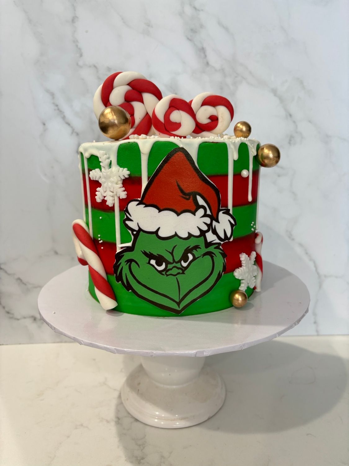 Grinch-themed Christmas cake on a white pedestal. Green, red, and white frosting with candy cane and Grinch's face.