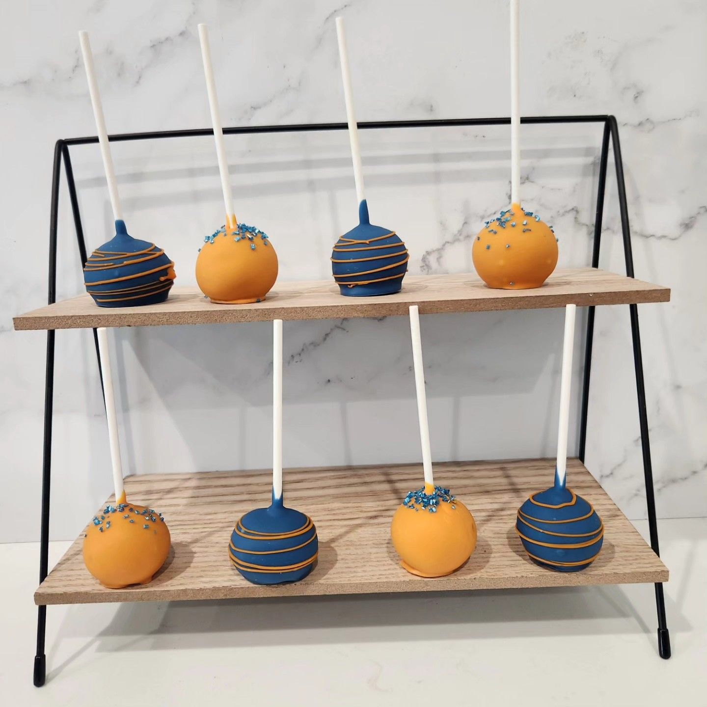 Cake pops on a two-tiered display. Navy blue and orange pops with gold detailing.