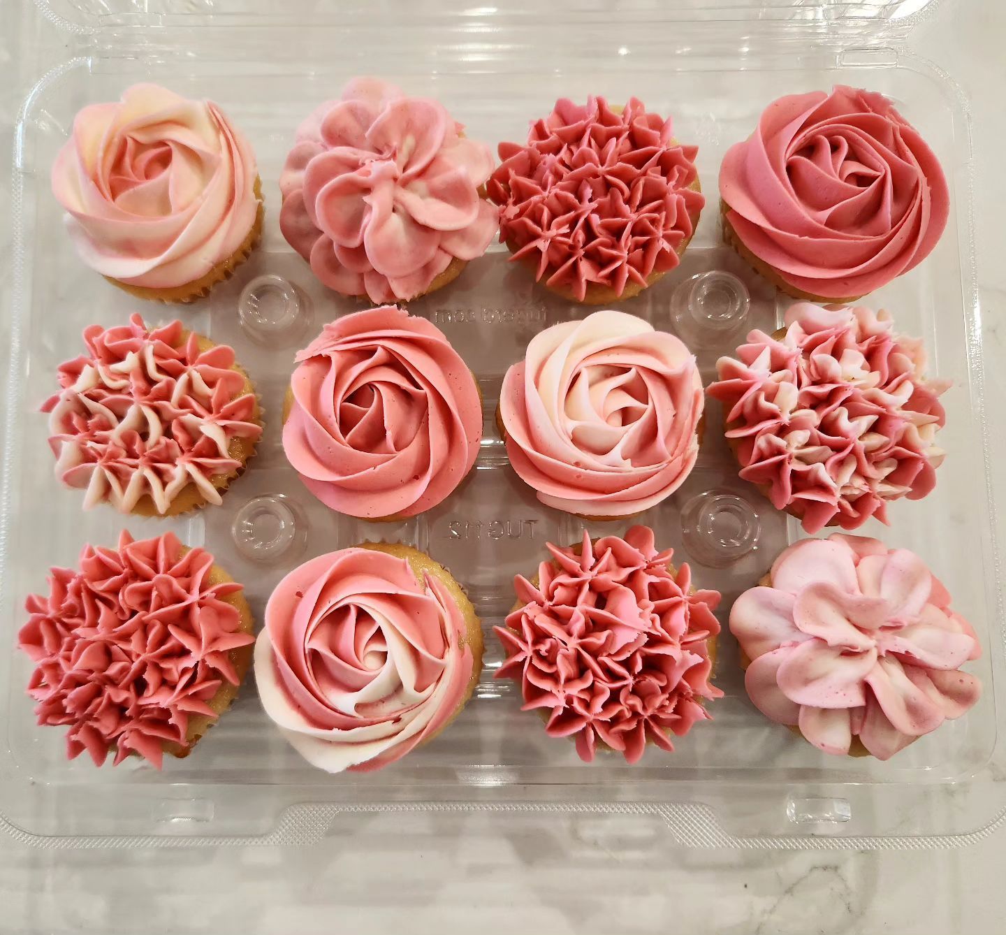 Twelve pink frosted cupcakes in a clear container, each with a unique floral design.