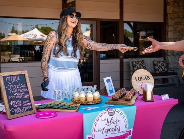 Woman with tattoos at a food stand, handing off a pastry. Pink table, baked goods.