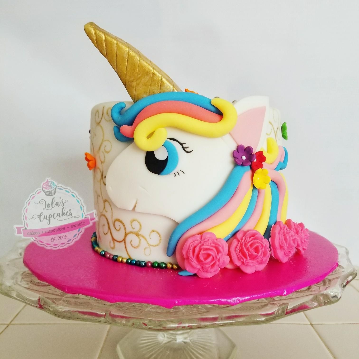 Unicorn cake with golden horn, colorful mane, and pink roses on a pink base, displayed on a glass cake stand.