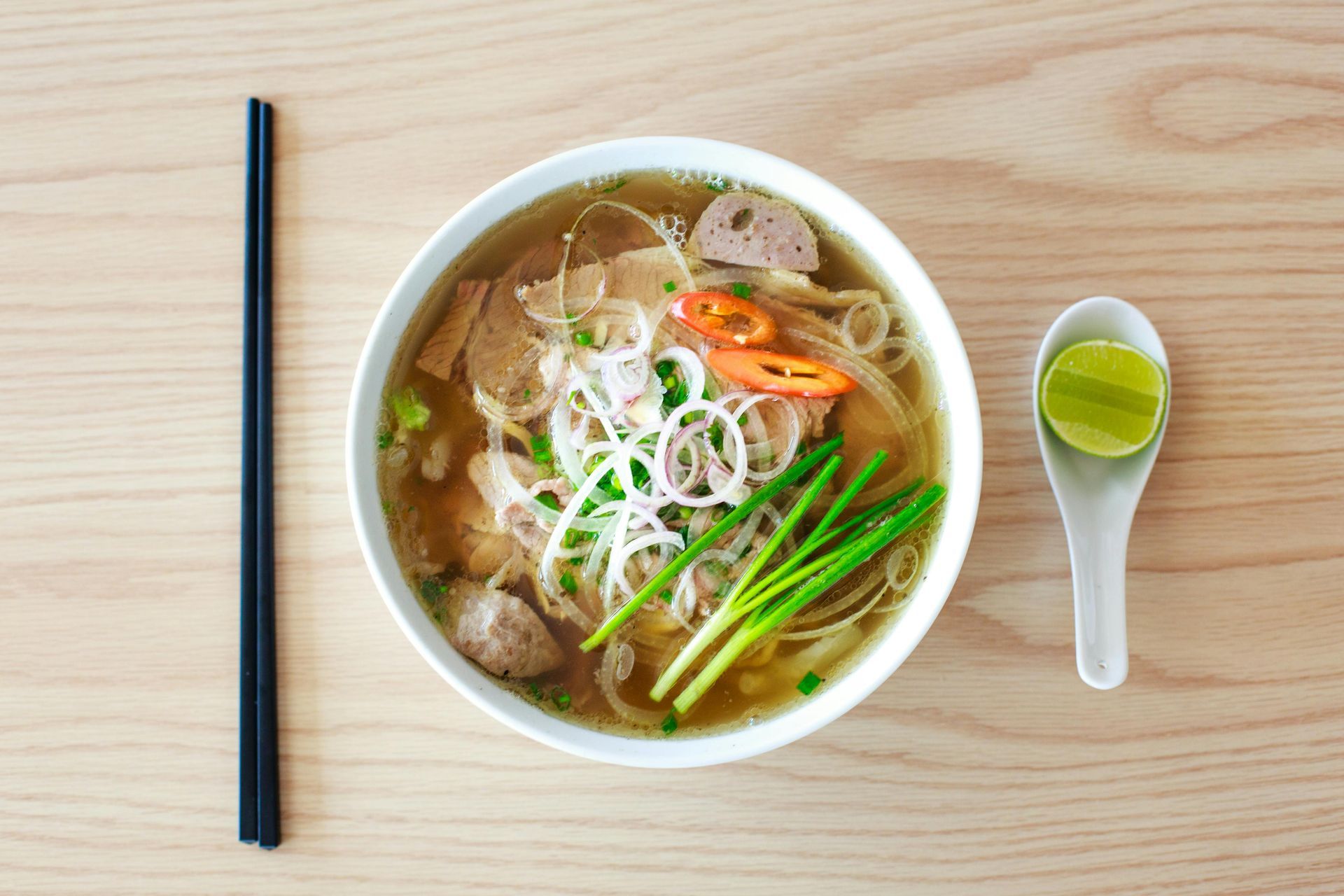 A bowl of soup with chopsticks and a spoon on a wooden table.