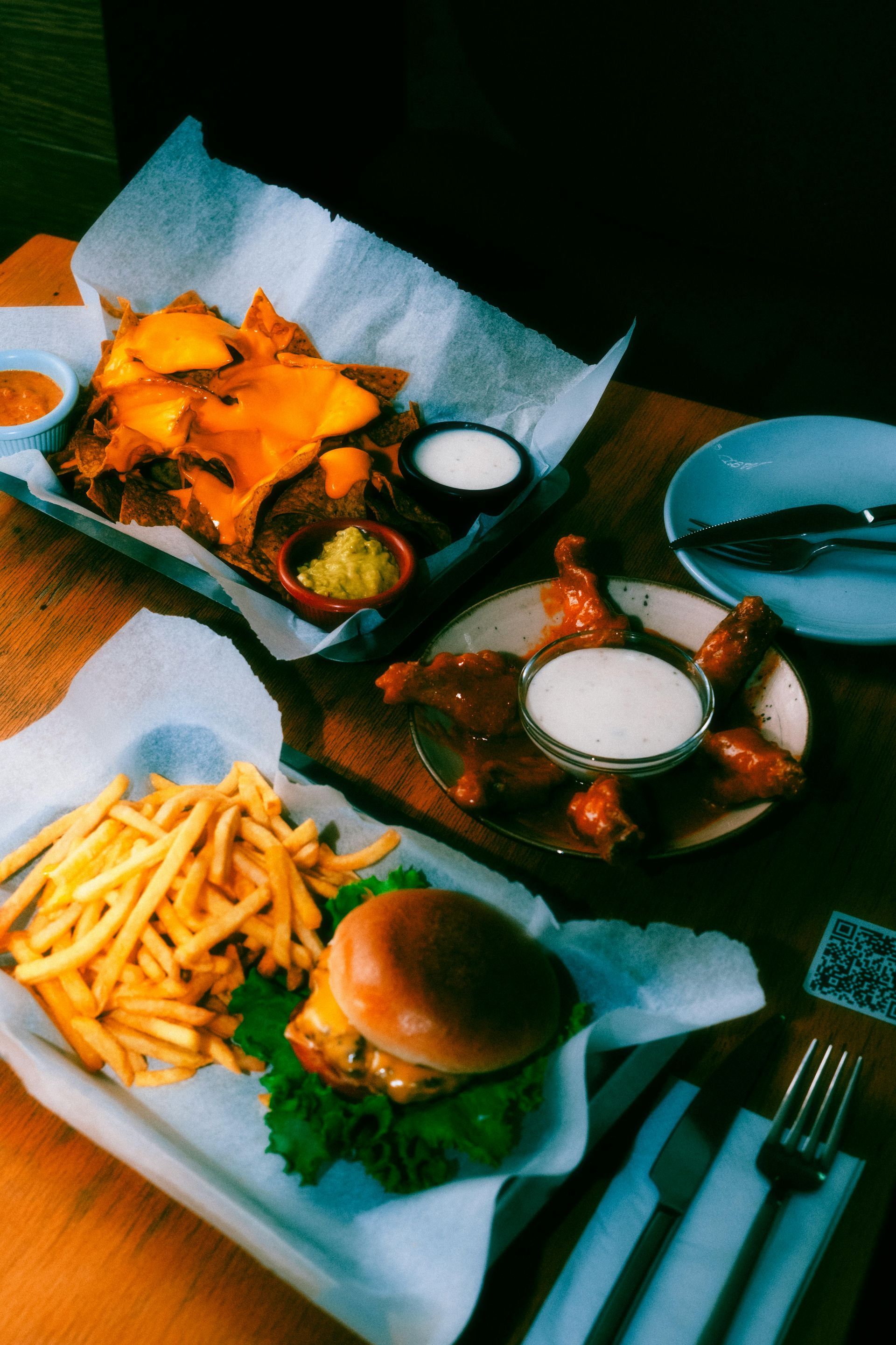 A table topped with plates of food including nachos , french fries , wings and a hamburger.