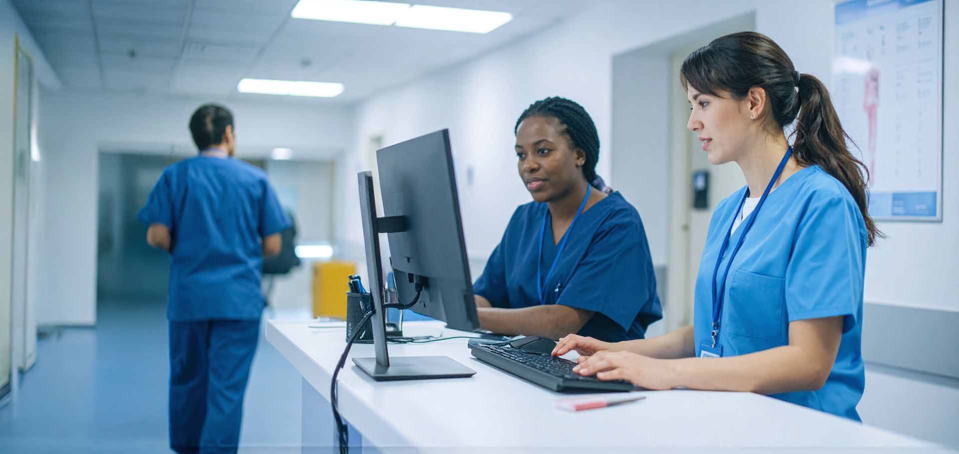 Two medical professionals looking at a computer in a hospital hallway. A third person is walking in the background.