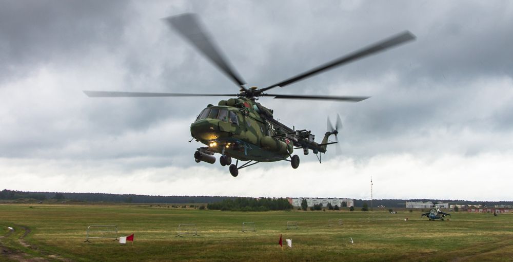 Military helicopter taking off from a grassy field under a cloudy sky.