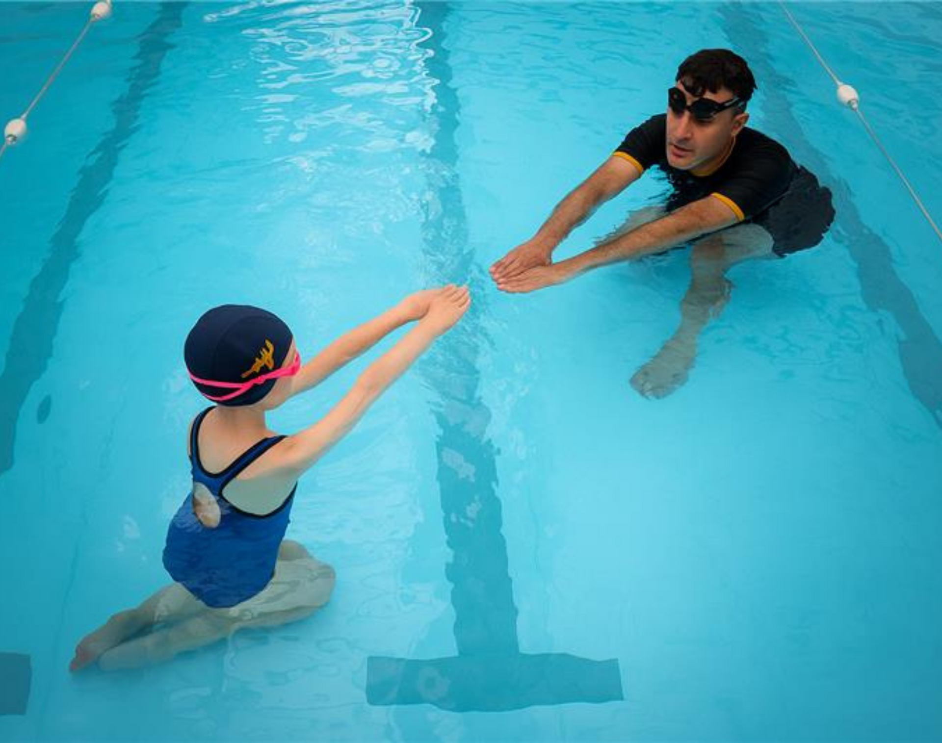 A child and instructor in a pool practicing swimming strokes, instructor demonstrating with arms outstretched.