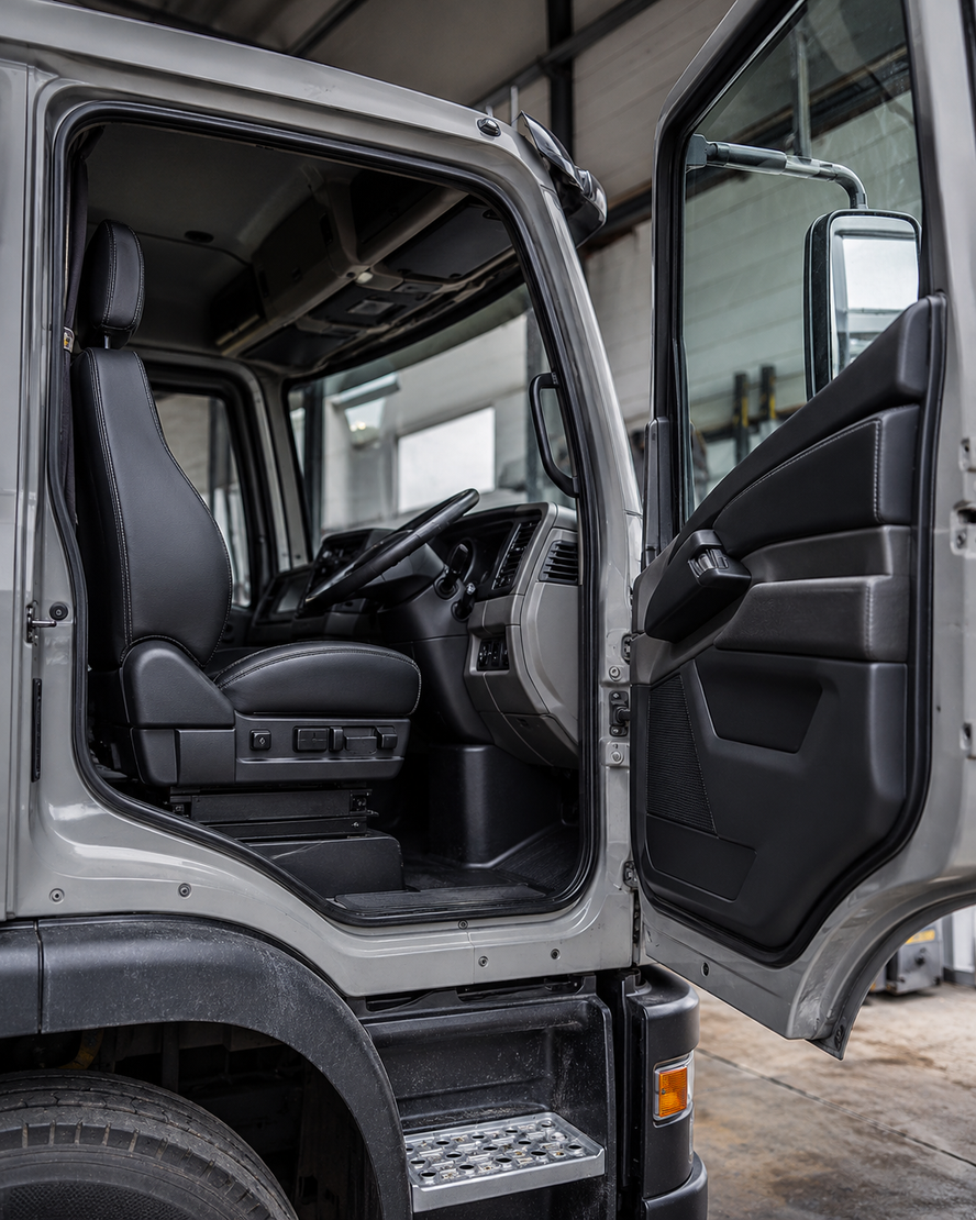 Cabina di camion aperta con portiera del conducente aperta, che mostra sedili neri e volante all'interno di un veicolo grigio.