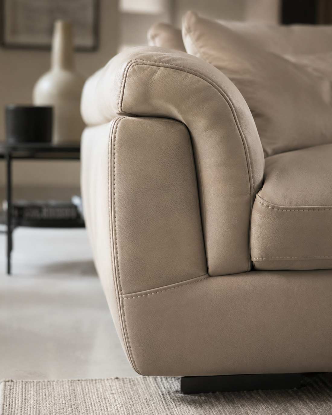 Bracciolo di divano rivestito in tessuto beige in un soggiorno moderno, con un tavolino e un vaso sfocati sullo sfondo.