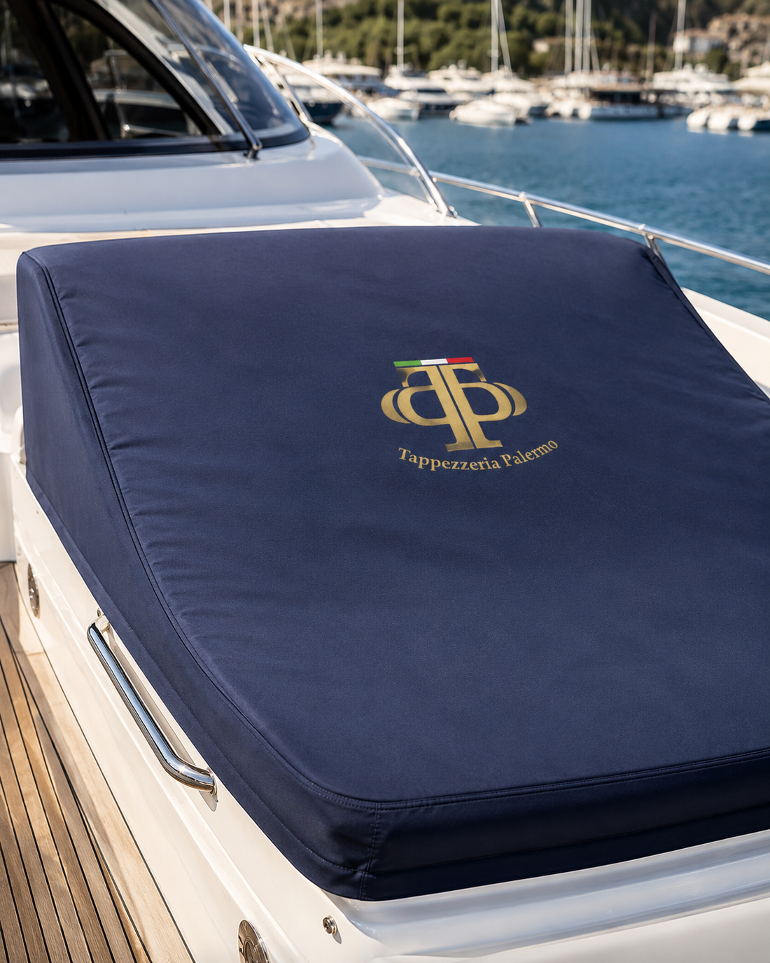 Cuscino blu da yacht con monogramma dorato sul ponte di una barca, con porto turistico e barche a vela sullo sfondo.
