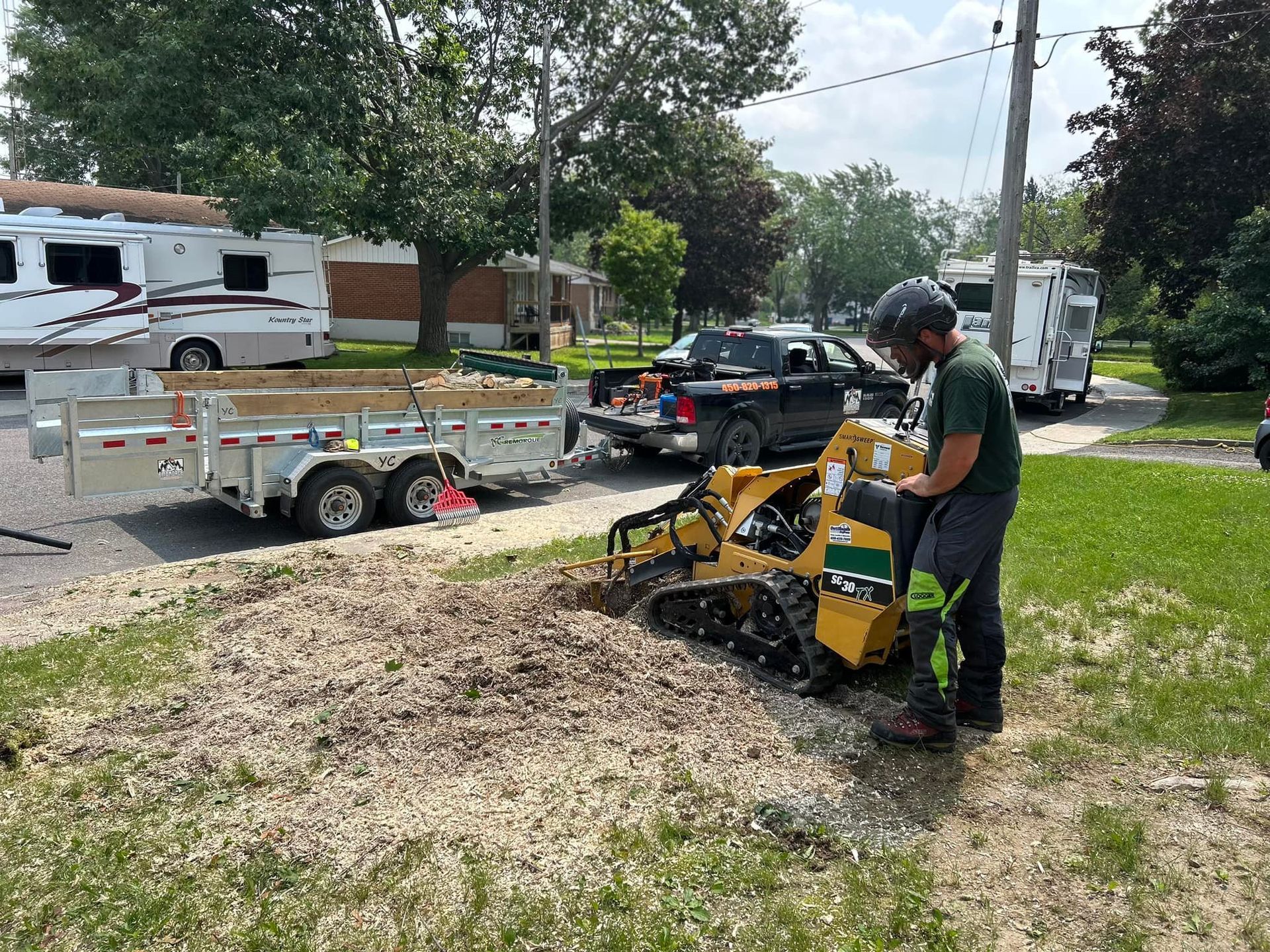 Person operating stump grinder, producing wood chips. Trailer, truck, and RVs visible. Outdoors, sunny.