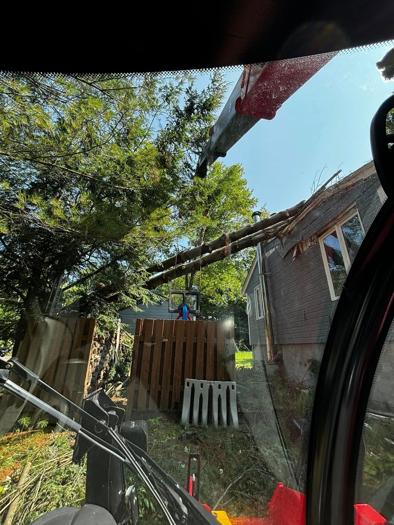 View from an excavator cab: beam lifted toward a house with tree and fence in the background.