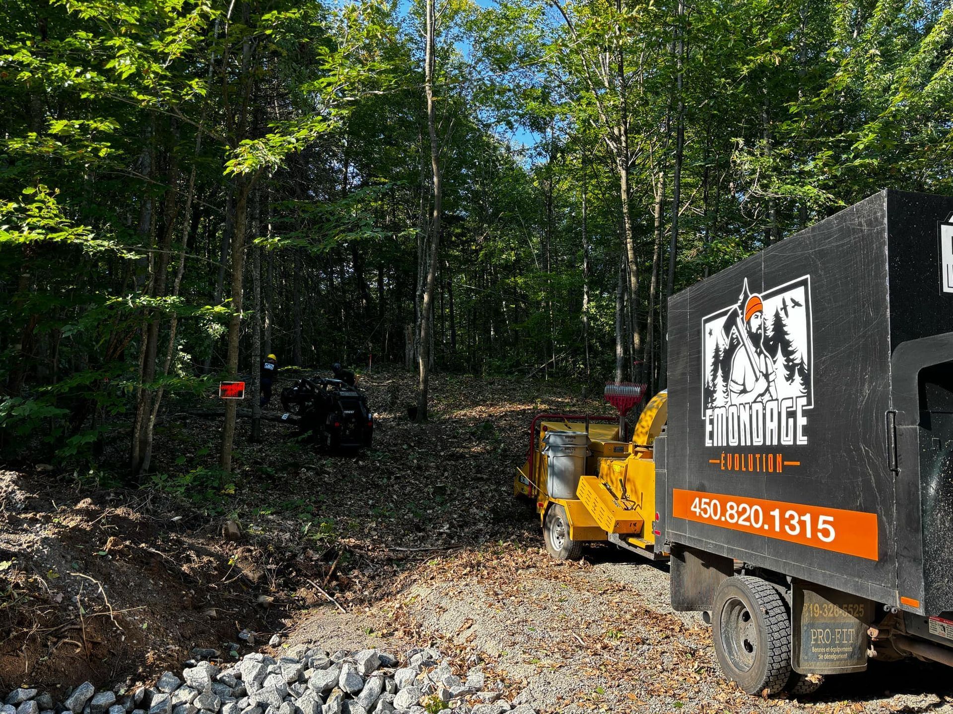 A wood chipper and truck in a wooded area, likely clearing debris. The truck has a logo and phone number.