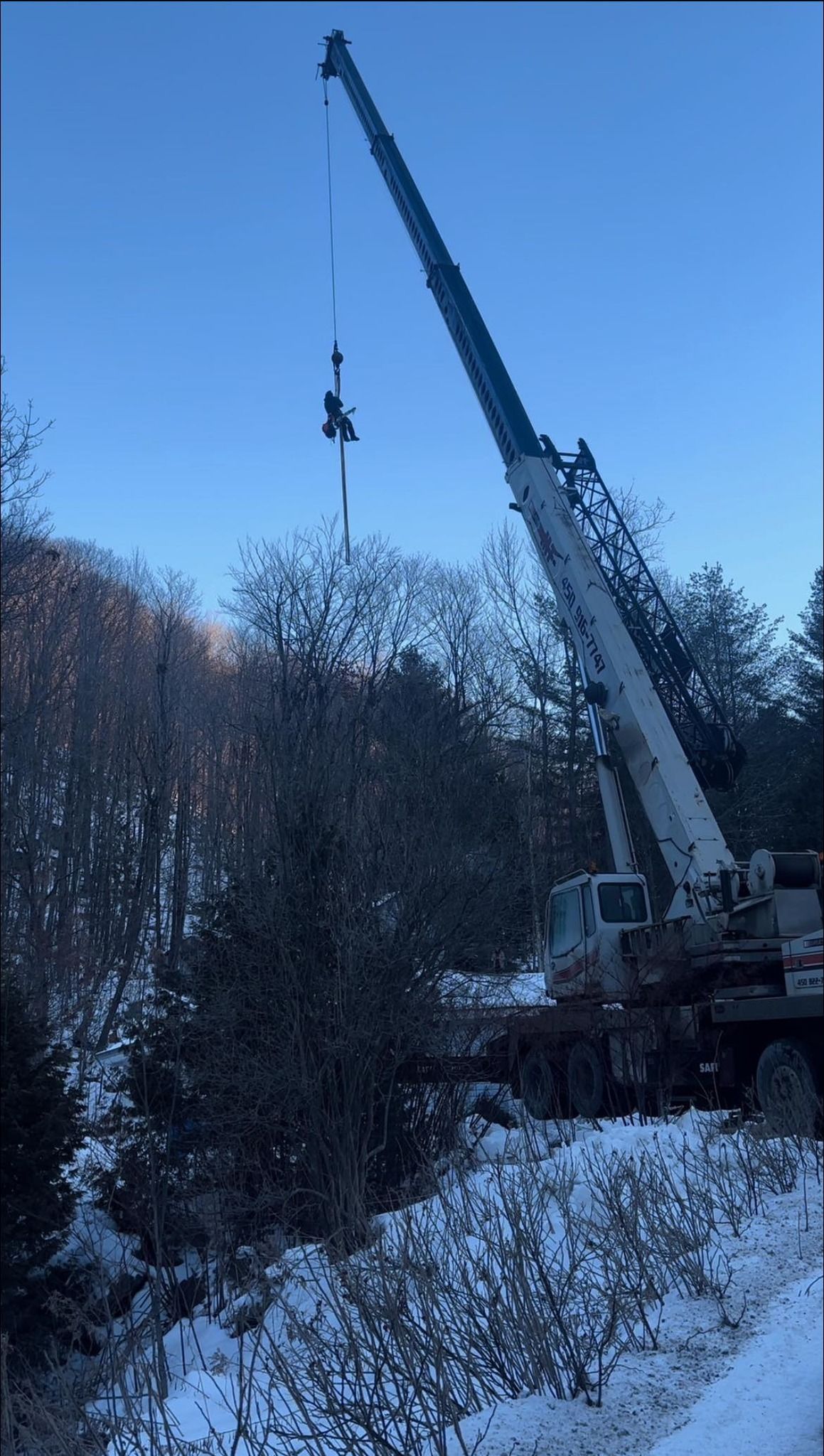 Crane lifting equipment over snow-covered trees and hillside on a clear day.