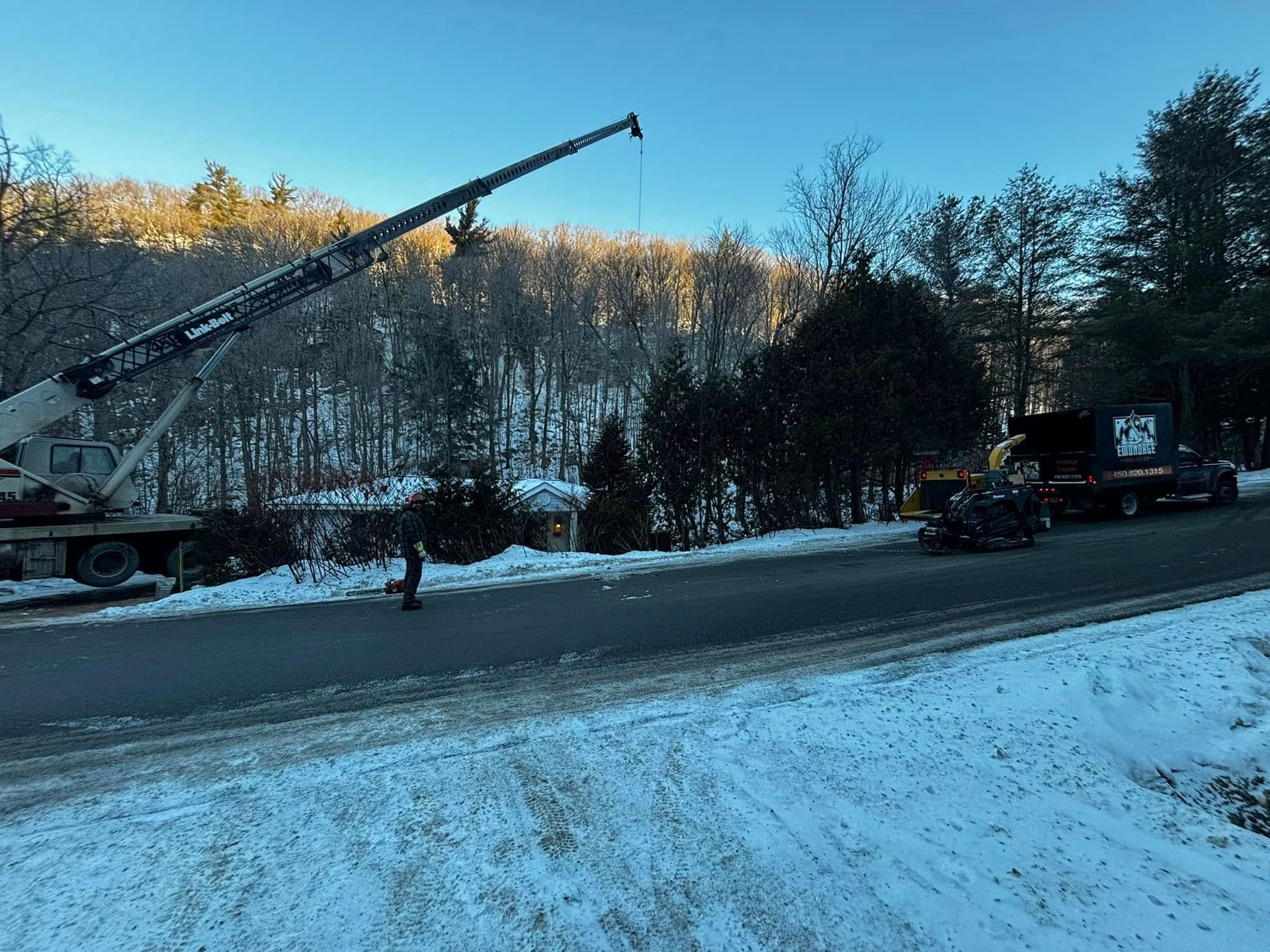 A crane lifting something near a snowy road, person walking, truck. Wooded background.