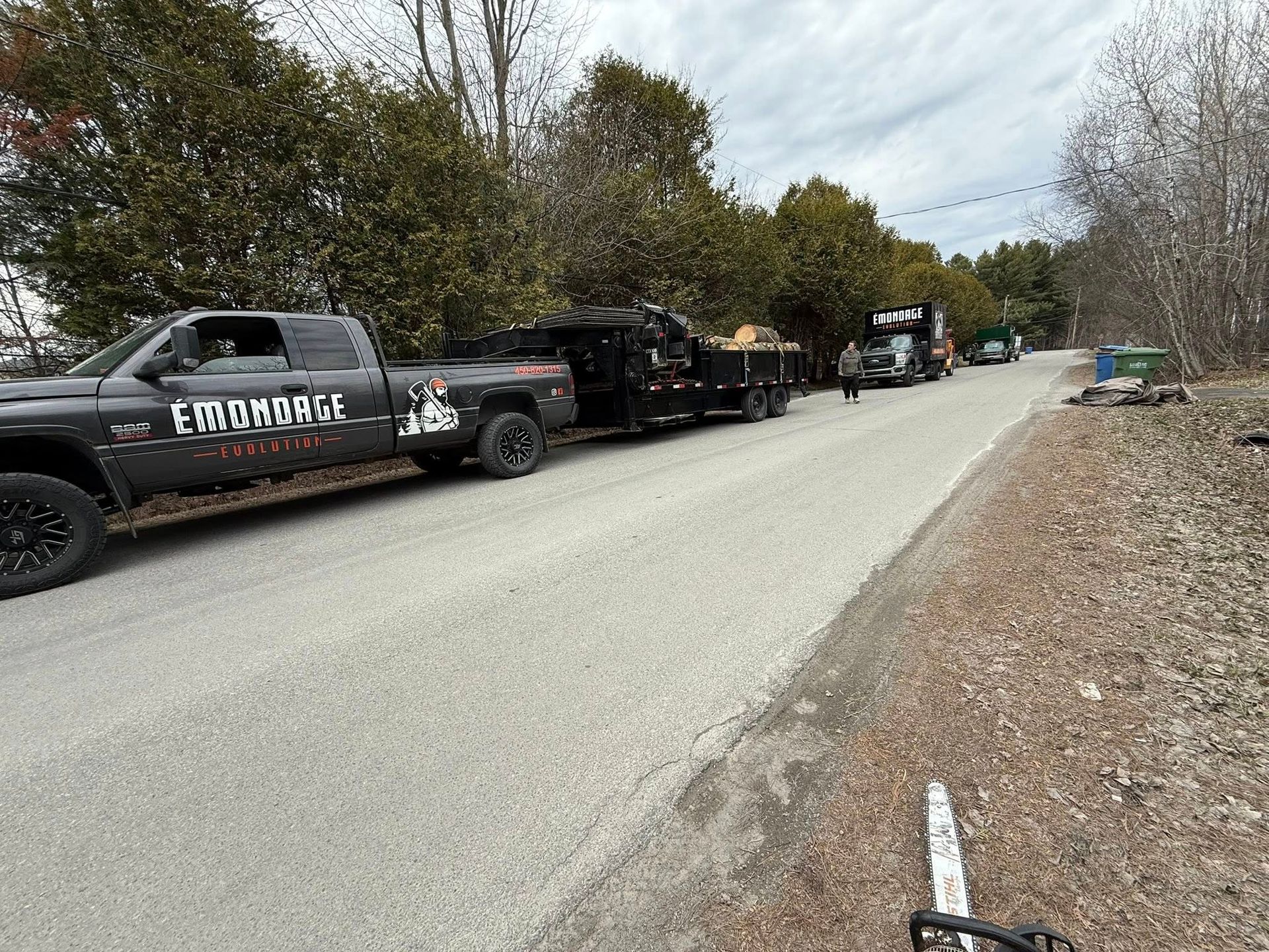 Truck with trailer on road, trees in background, person walking, chainsaw in foreground.