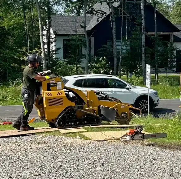 Man operating a yellow stump grinder next to a white SUV. A chainsaw sits on the ground.
