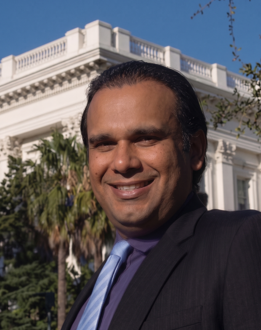 Smiling man in a dark suit standing outdoors before a white columned building.