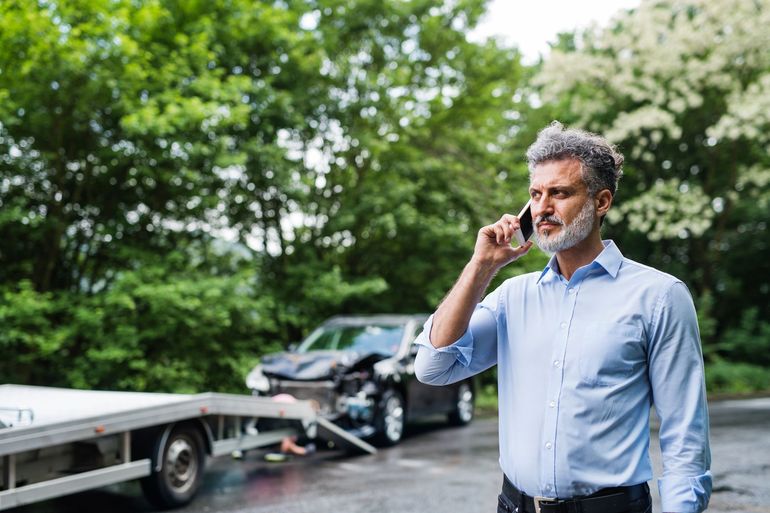 Man in a blue shirt on a phone beside a tow truck and damaged car on a roadside