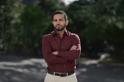 Man standing outdoors with arms crossed, wearing a maroon shirt and light pants, against a blurred background