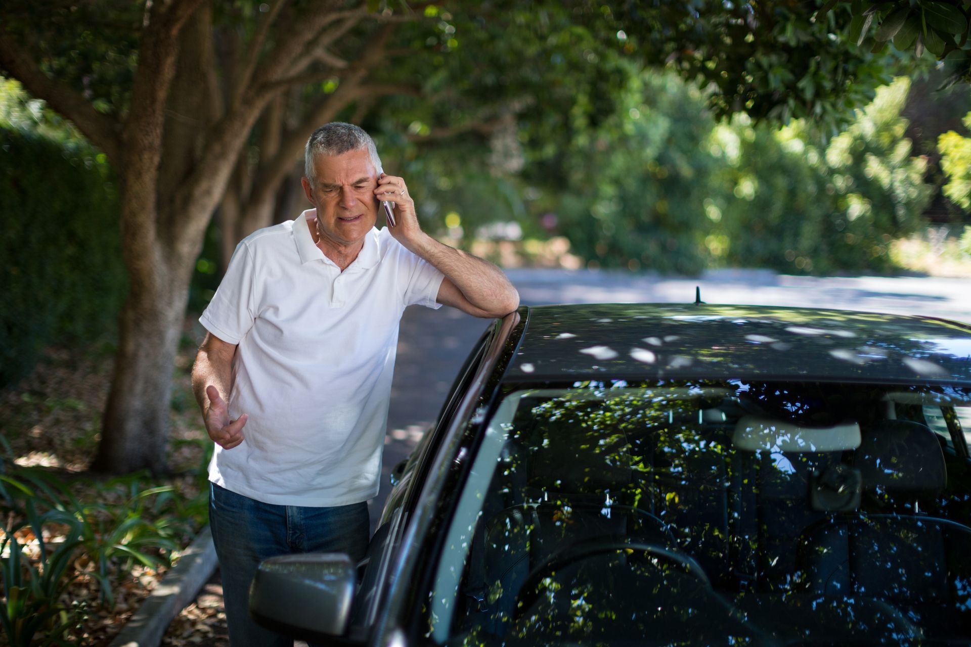 Man leaning on a black car under trees, speaking on a mobile phone outdoors