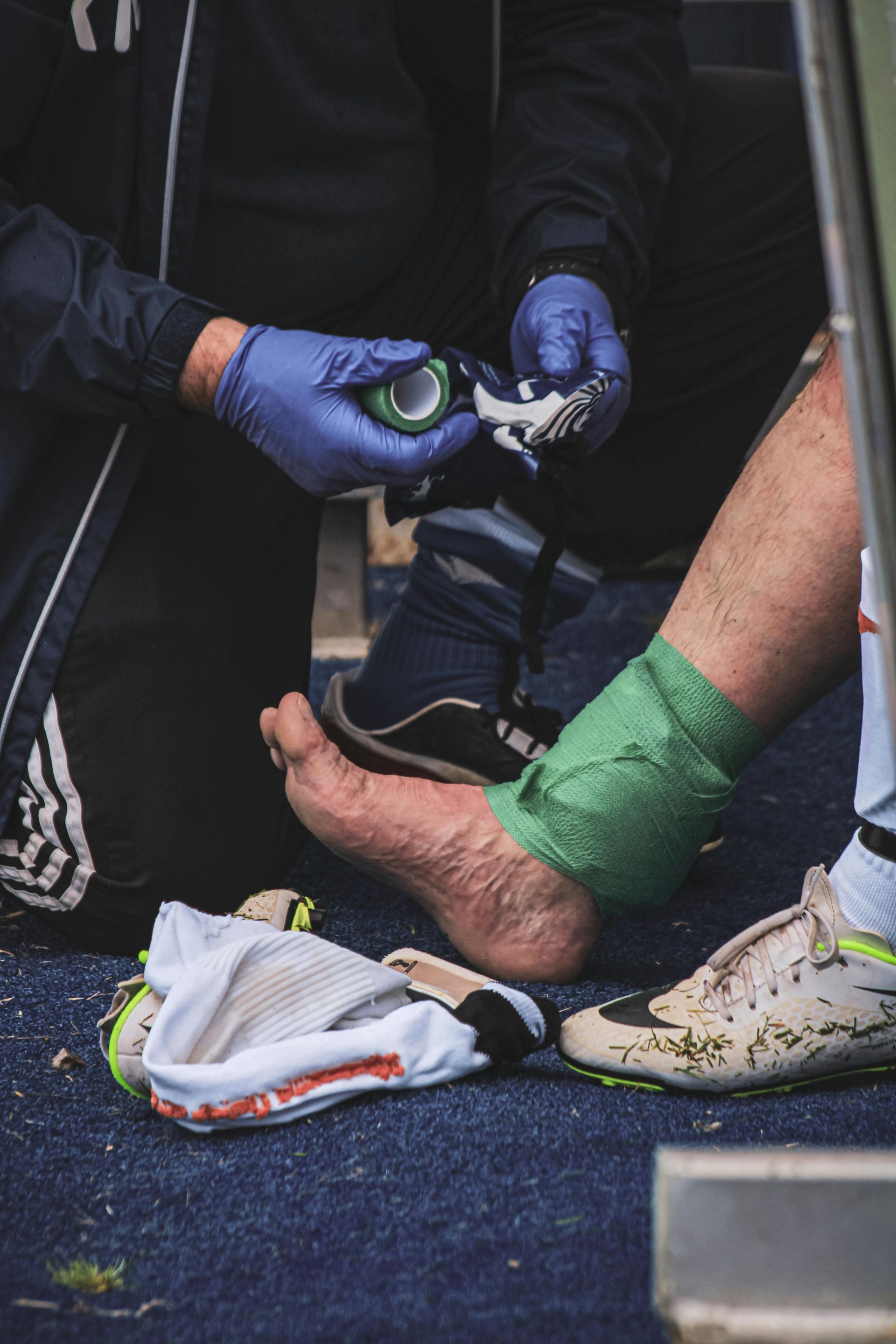Gloved hands bandaging a bare foot with a green wrap on a dark floor