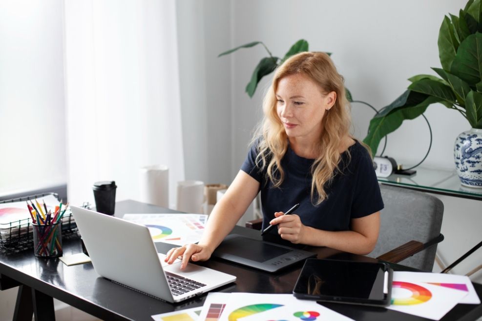 Woman using laptop and tablet at desk with color palettes and plants.