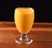 Mango smoothie in a stemmed glass on a wooden surface against a black background.