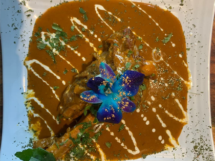 Chicken curry in a brown sauce, drizzled with cream, garnished with herbs and a blue flower, on a white plate.