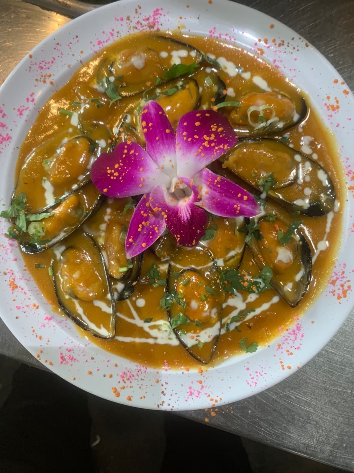 Mussels in orange sauce with cream drizzle, garnished with an orchid and herbs on a white plate.