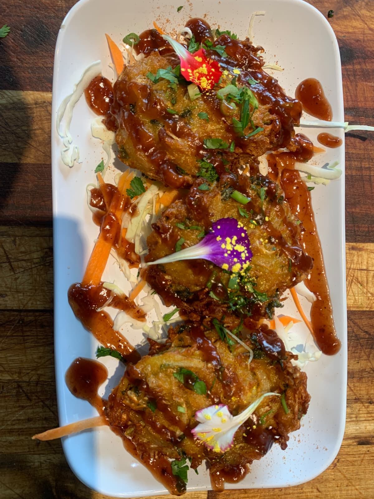 Three fried food patties drizzled with sauce, garnished with flowers and herbs on a bed of shredded vegetables.