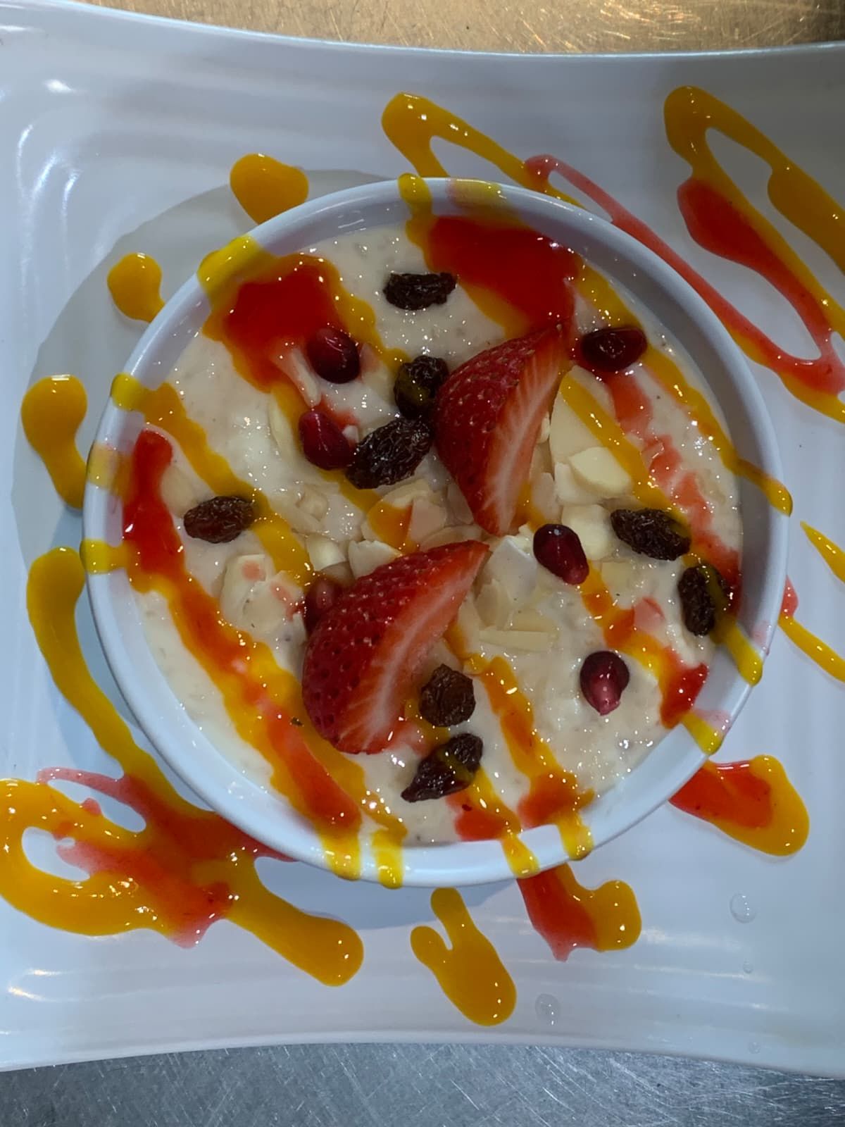 Dessert bowl with creamy pudding, strawberries, and raisins, drizzled with orange and red sauces.