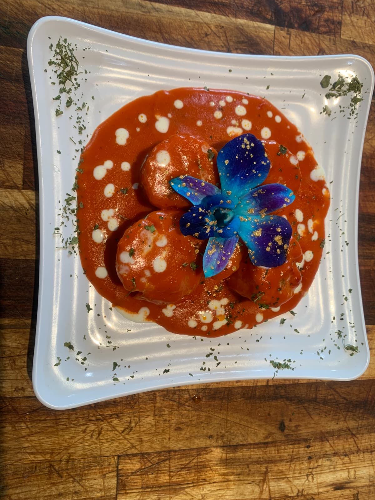 Meatballs in a red sauce, dotted with white, garnished with a blue flower on a white plate, sprinkled parsley.