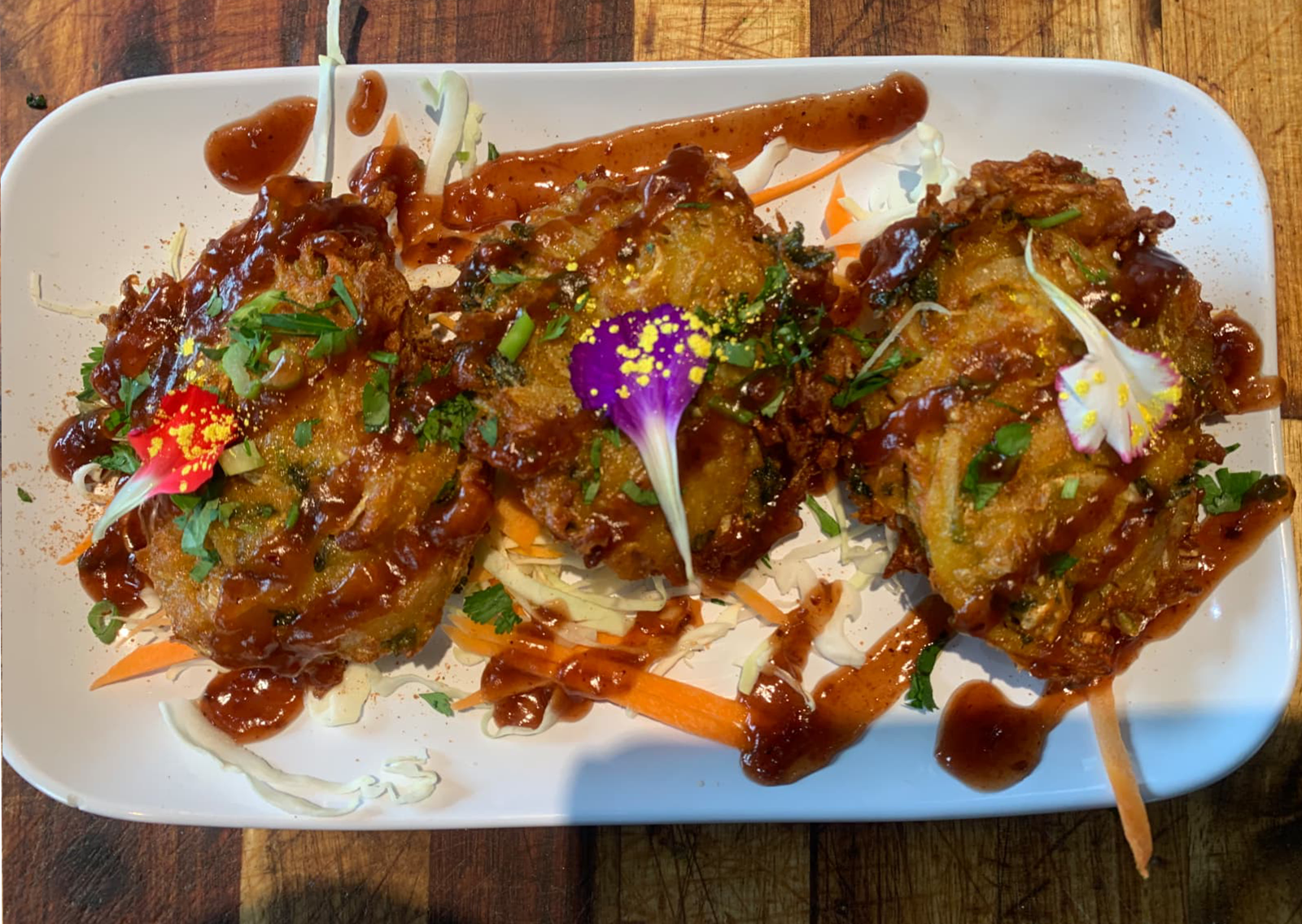 Three fried savory patties drizzled with sauce on a white plate with carrot and cabbage garnish, topped with flowers.