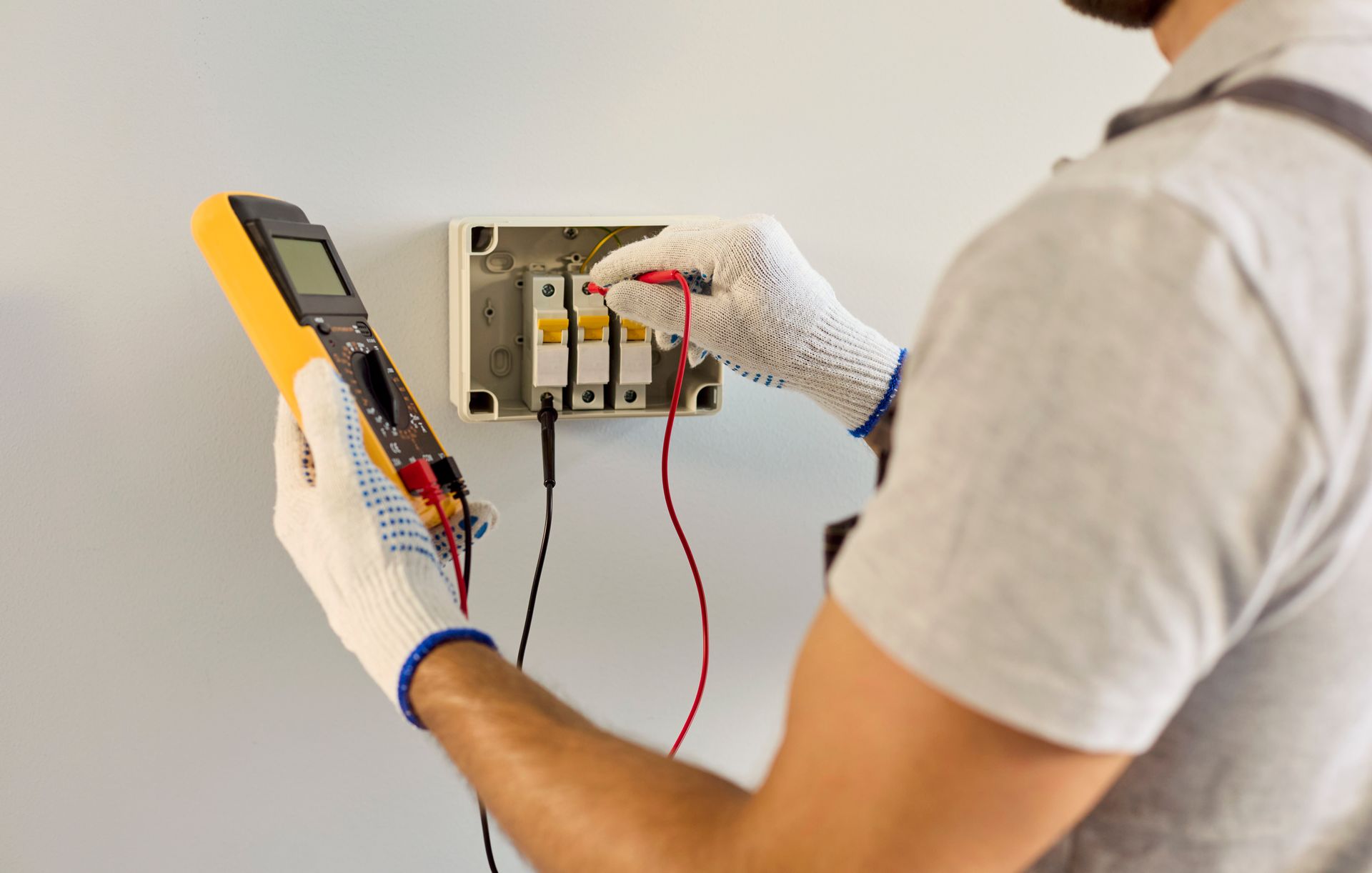 Electrician testing wall wiring with a multimeter, checking an open electrical panel.