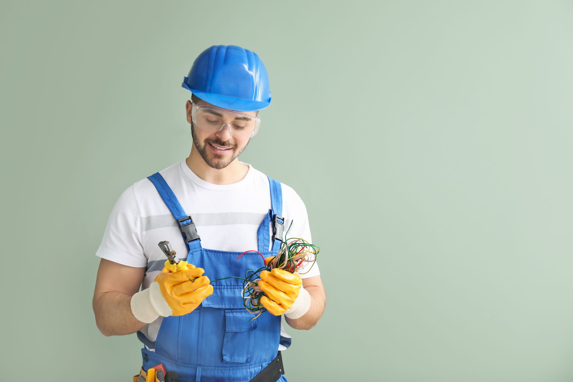 Worker in blue overalls and hard hat holding wires and tools against a plain green background