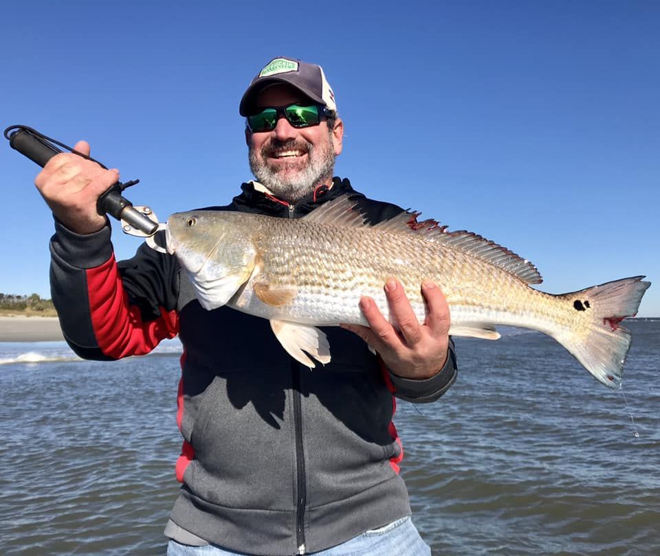 Smiling angler holding a large striped fish on a sunny beach with ocean waves behind him