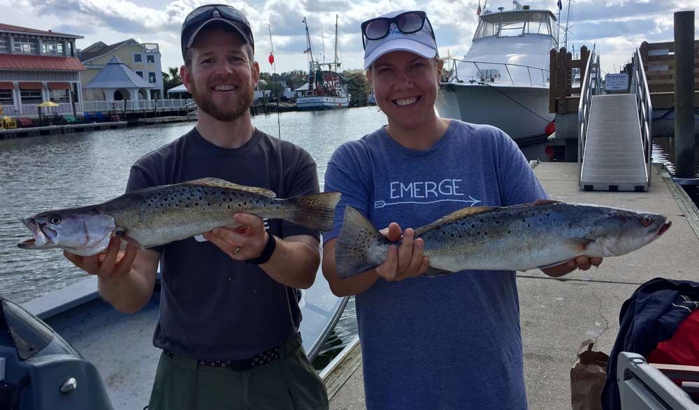 Two men on a dock holding large fish, smiling with a fishing boat in the background.